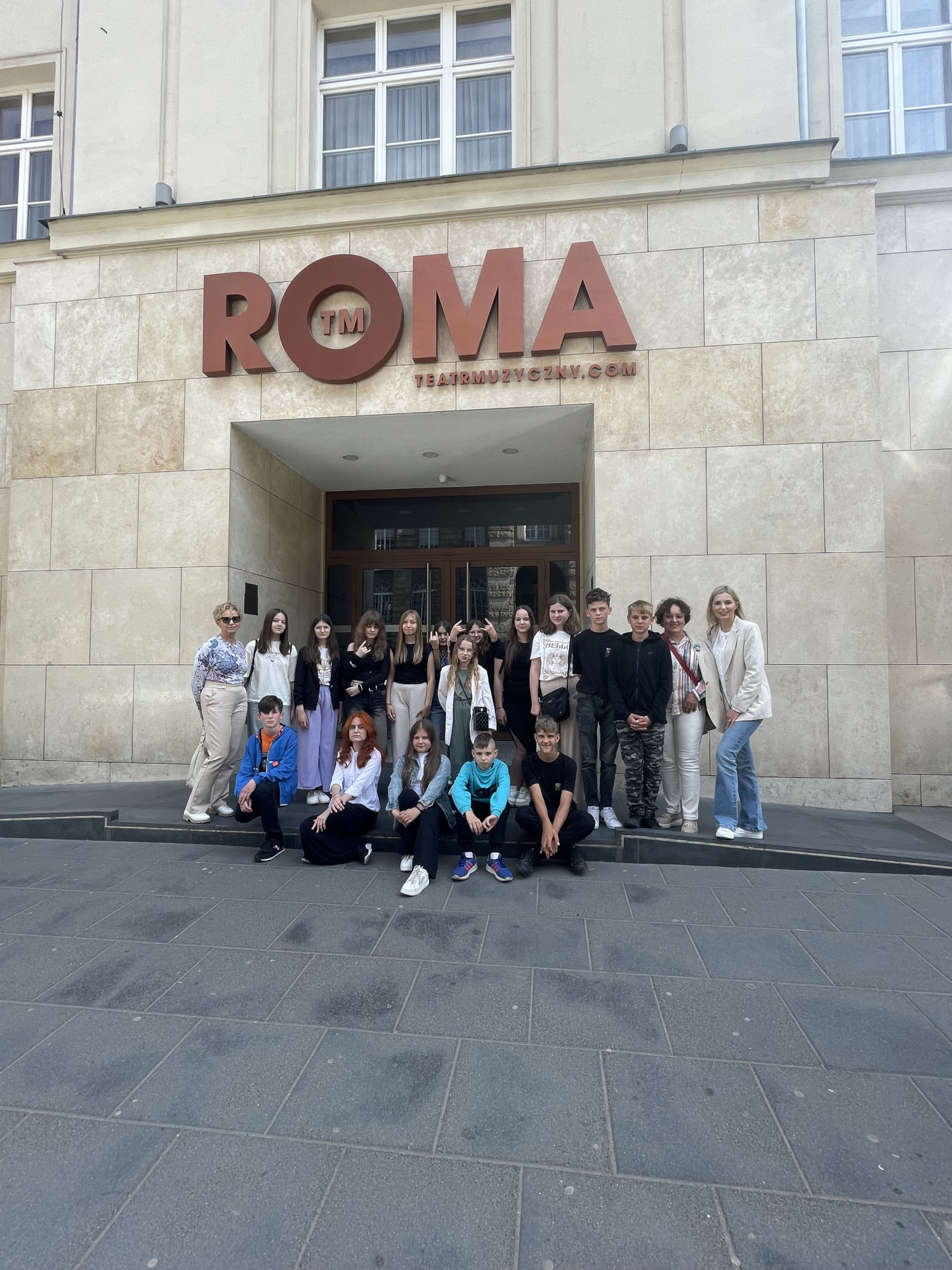 Grupa osób na tle budynku teatru Roma. Młodzież szkolna oraz nauczyciele.