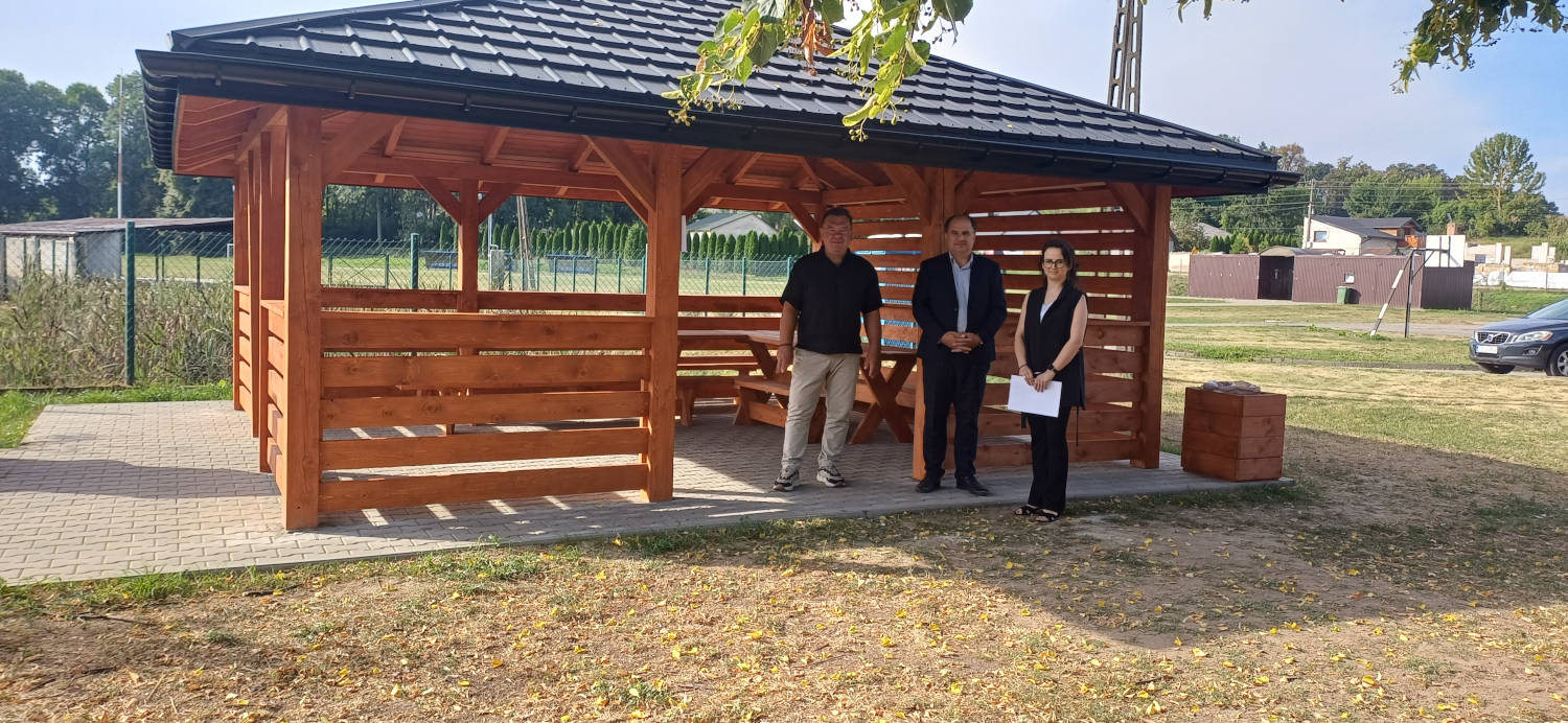 Odbiór altanki z wyposażeniem przy stadionie sportowym w Wojciechowicach Wójt oraz dwoje pracowników Urzędu Gminy na tle drewnianej altany