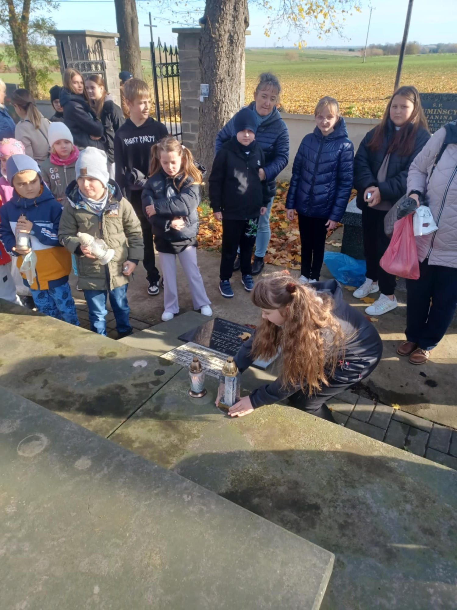 Zdjęcie 2 Uczniowie wraz z nauczycielką przed mogiłą, dziewczynka ustawia znicz na pomniku