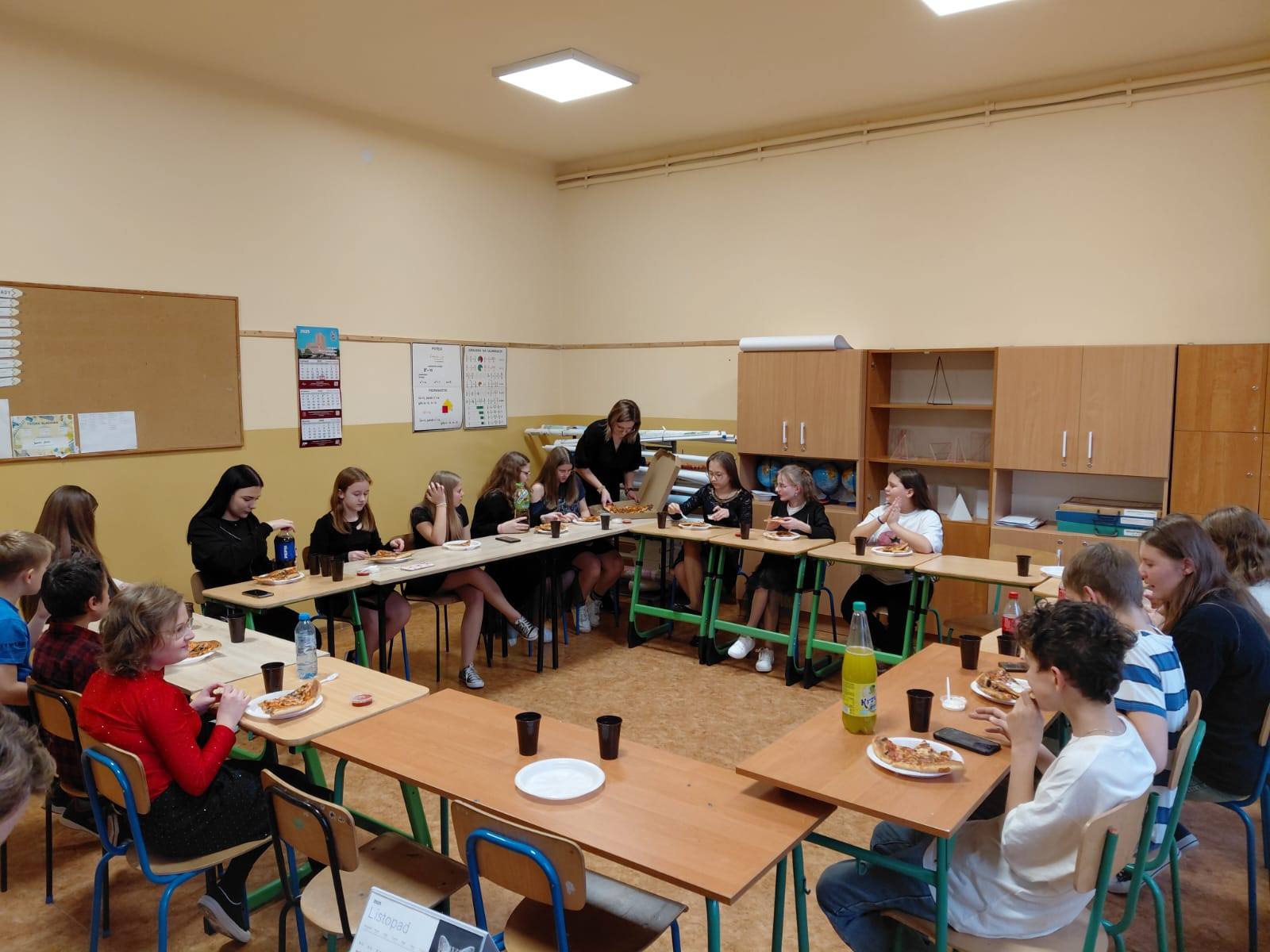 Uczniowie siedzą w ławkach ustawionych w kwadrat i jedzą pizze