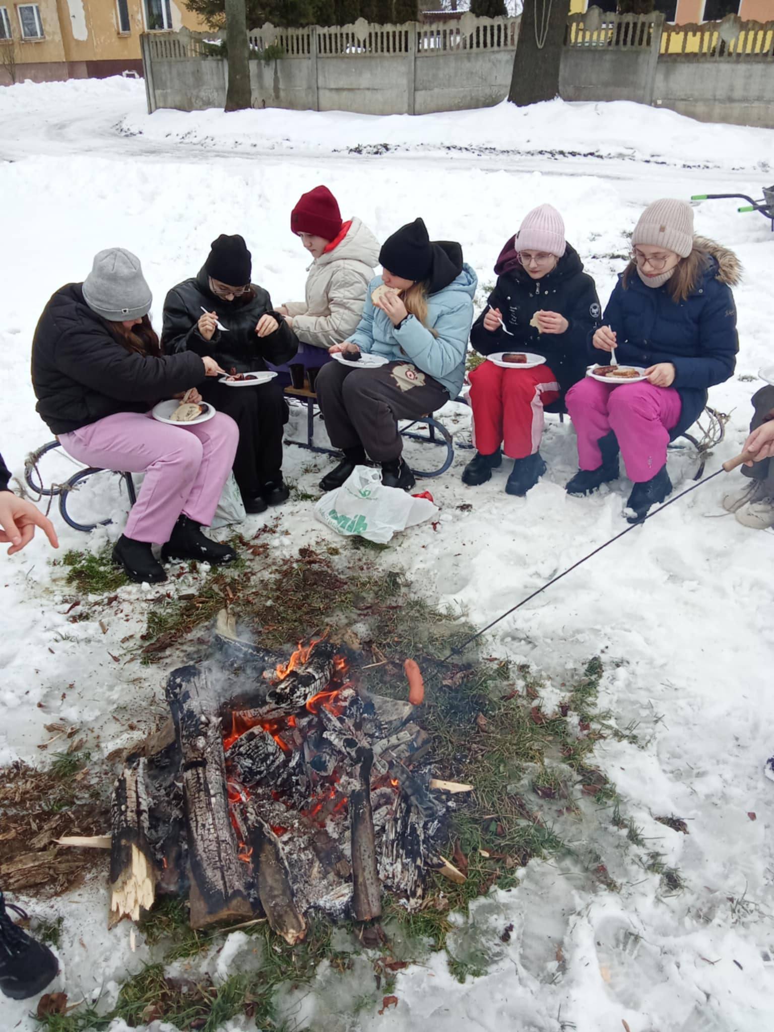 Dzieci jedzą kiełbaski siedząc na sankach przy ognisku